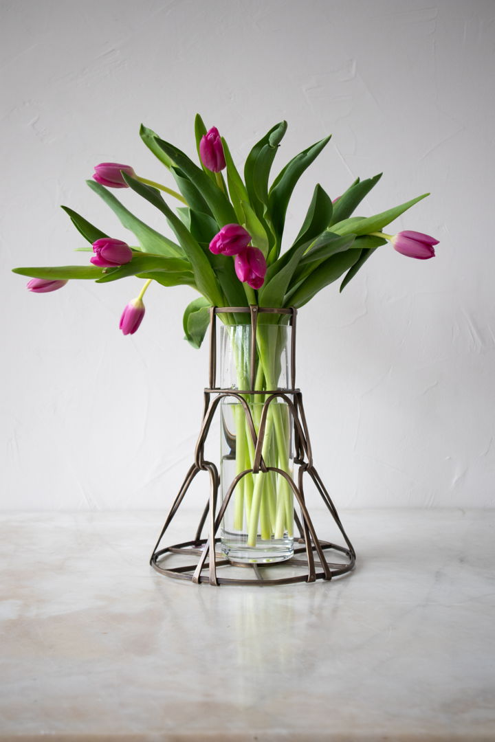 This basket vase features a woven design made from forged brass. It can be used to elevate a bouquet of fresh flowers  or simply as a one-of-a-kind artistic piece. A glass cylinder is also included. Shown in antique brass patina.