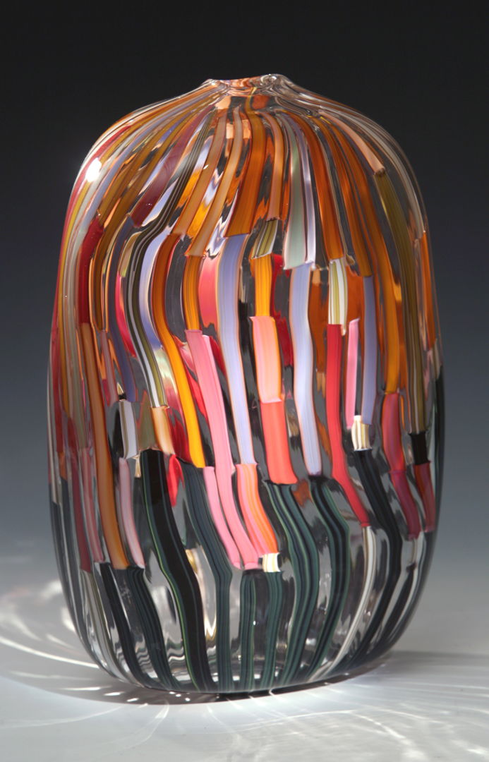 Blown glass form with multicolored cane work arranged in a rising dark to light pattern. This piece is shaped into a rounded rectangular form.