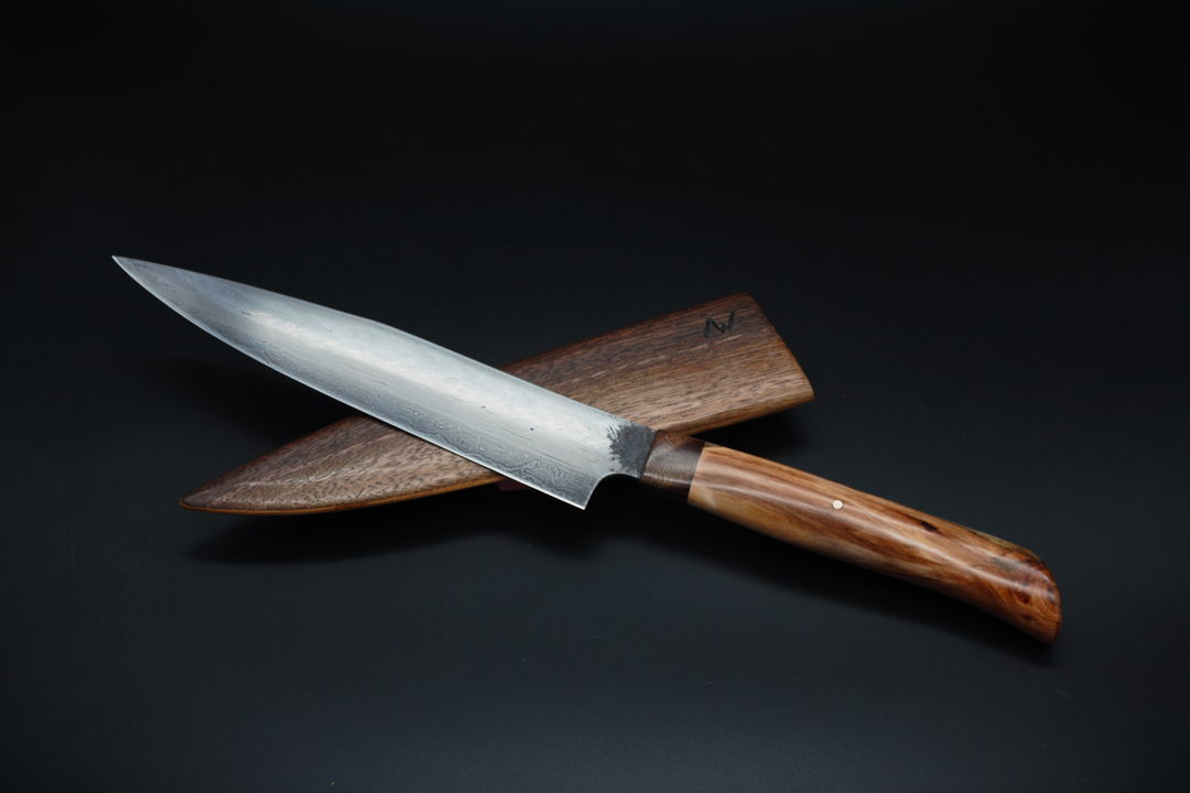 Forged from more than fifty bars of bloomery steel  this chef knife's 8" blade is decorated with the water ripples and herringbone twists that adorned ancient sword blades. The subtly patterned blade is mounted in a handle carved from stabilized walnut and apple wood  and can be sheathed in a walnut scabbard.