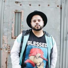 Man with a beard and a black hat stands outside in front of a metal door wearing a tee shit that says Blessed
